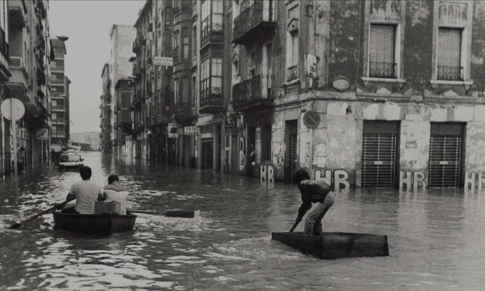 Imagen en blanco y negro donde aparecen edificación antiguos. El agua inunda las calles, alcanzando unos 50 cm. De altura. Varias personas se desplazan por el agua en improvisadas barcas creadas con maderas. 